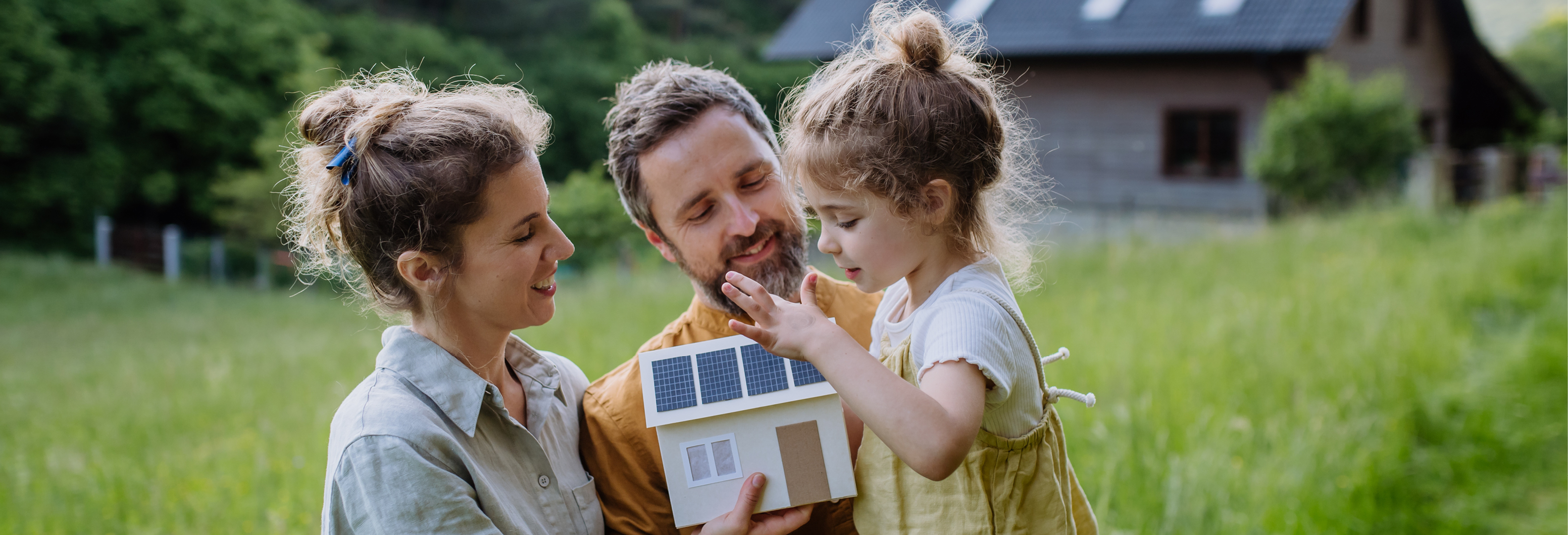 Eine Personengruppe steht auf einer Wiese und hält ein Modellhaus mit Solarpaneelen, im Hintergrund ein Haus mit Solardach.